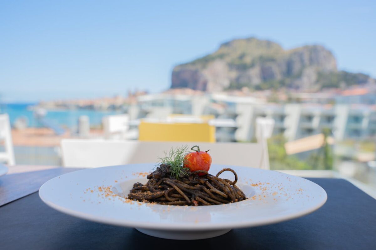 heros-img Plate of squid ink pasta with tomato and herbs, set against a beautiful seaside view.